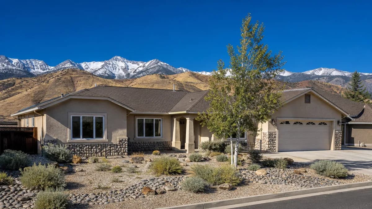 Single-story tan stucco ranch home with white vinyl windows, a xeriscaped front yard of sagebrush and river rock, and snow-dusted Sierra peaks behind